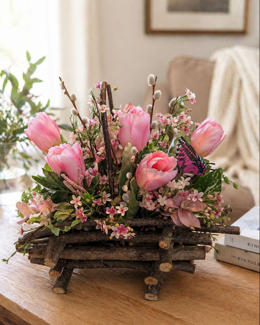 Decorative floral arrangement with pink tulips and a butterfly on a wooden base, placed on a table.